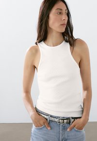 Hvid ribbet tanktop med høj hals, kombineret med lyseblå denimjeans og et dekorativt bælte med perleaccent.