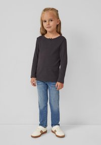 Mädchen steht vor einem schlichten grauen Hintergrund und trägt ein dunkles Langarmshirt, blaue Jeans und weiße Sneakers mit braunen Akzenten.