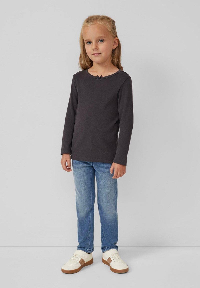 Mädchen steht vor einem schlichten grauen Hintergrund und trägt ein dunkles Langarmshirt, blaue Jeans und weiße Sneakers mit braunen Akzenten.