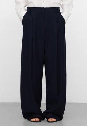 Pantalon large bleu marine avec plis et poches latérales, confectionné dans un tissu léger. L'ourlet tombe librement sur des sandales noires.