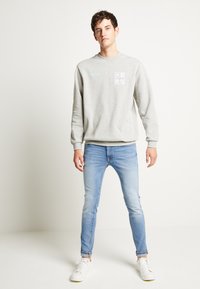 Sweatshirt cinza com detalhes impressos e jeans azuis, complementados por ténis brancos. Design simples e corte relaxado.