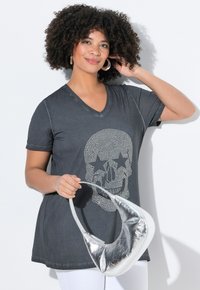 Grå V-hals t-shirt med rhinsten-skalle-grafik, kombineret med en metalisk sølv taske. Modellen har krøllet hår og holder tasken.