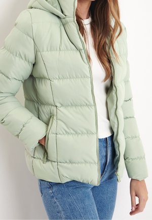 Femme portant une veste matelassée à capuche vert clair sur un haut blanc et un jean bleu, avec une main dans la poche de la veste.