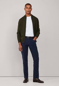 Tamnozeleni kardigan preko bijele košulje, tamnoplave hlače i smeđe cipele. Kardigan ima zatvarač na patent i rebrastu teksturu.