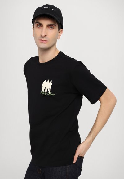 Melnas kokvilnas T-krekls ar īsām piedurknēm, uz kura ir neliela grafika ar trim figūrām un teksts "Makšķerēšanas gabali" zaļā krāsā. Modelis nēsā melnu cepuri.