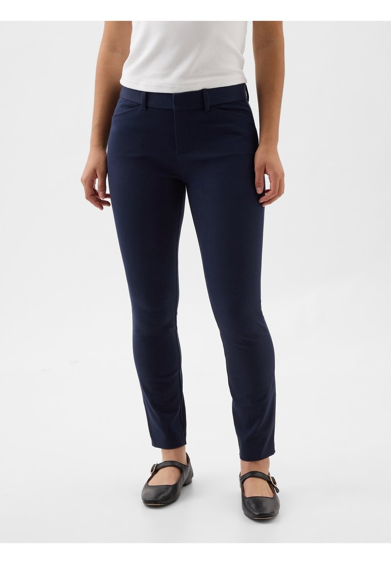 Personne portant un pantalon bleu marine ajusté, un haut blanc et des chaussures Mary Jane noires, debout devant un fond blanc uni.