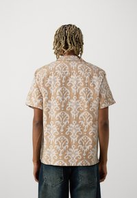 Camicia a maniche corte beige con motivo bianco ornamentale, dotata di colletto, vestibilità comoda e texture morbida.