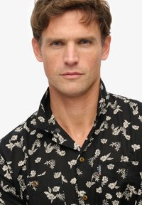 Camisa negra de manga corta con un patrón floral en crema, con cuello y botones marrones. Incluye un bolsillo en el pecho con una etiqueta.