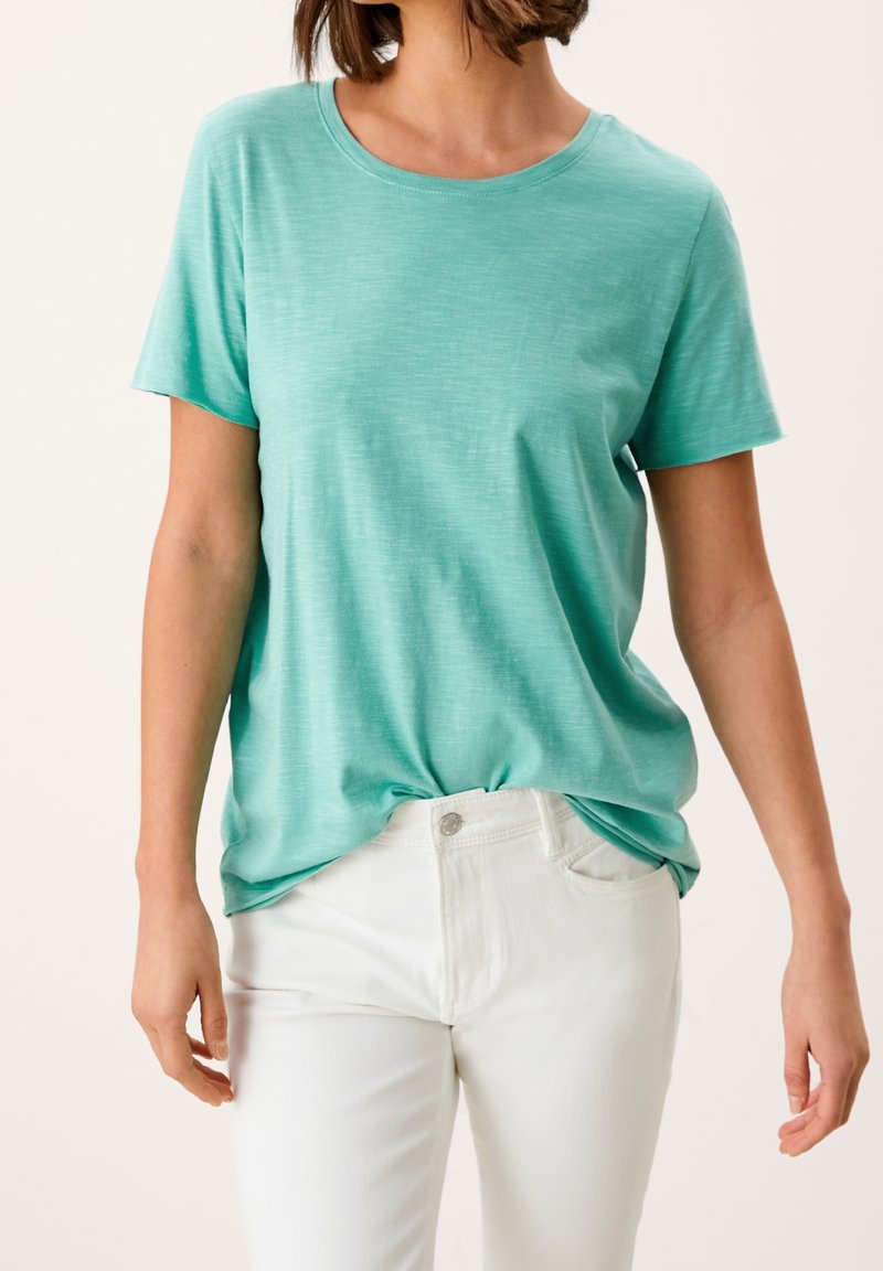 Femme portant un t-shirt à manches courtes bleu sarcelle clair légèrement rentré dans un pantalon blanc sur un fond uni.