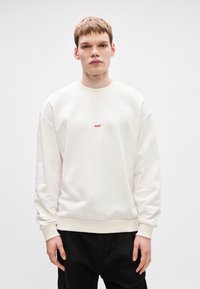 Ανοιχτό μπεζ φούτερ σε χαλαρή γραμμή, με στρογγυλή λαιμόκοψη και ραμμένες μανσέτες, διαθέτει ένα μικρό κόκκινο λογότυπο "HUGO" στο κέντρο.