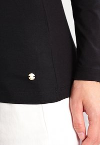 Schwarzes Langarmshirt mit einem runden, goldfarbenen Metalldetails mit dem Markennamen nahe dem Saum, aus weichem, dehnbarem Material gefertigt.