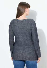 Žena s dlouhými hnědými vlasy, nosící stříbrné kruhové náušnice, námořnický modrý pruhovaný svetr s dlouhým rukávem a modré džíny, obrácená zády.