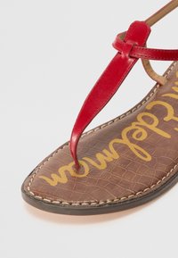 Red leather thong sandal with a brown textured insole featuring raised patterns and yellow branding. Stitched detailing around the sole.