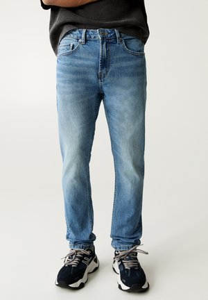 Light blue denim jeans with a straight-leg fit, medium wash, five-pocket design, and a button fly. Worn with black and white sneakers.