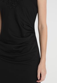 Robe noire sans manches avec des détails en dentelle texturée au niveau de l'encolure. Tissu lisse qui tombe doucement sur la silhouette. Coupe ajustée.