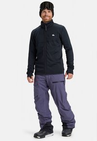 Men's black zip-up jacket with high collar, paired with loose-fitting purple snow pants, featuring multiple pockets and black accents.