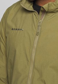 Olivově zelená zipová bunda vyrobená z měkkého materiálu, s vysokým límcem, viditelným zipem a stříbrným logem "Mammut" na levé části hrudi.
