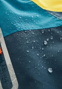 Waterdichte stof in blauw, geel en donkergrijs met zichtbare waterdruppels. Glad oppervlak met een logodetail en levendige kleurcontrasten.
