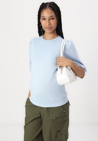 Suéter azul claro de mangas curtas com mangas bufantes, combinado com calças cargo verde-oliva e uma pequena mala branca.