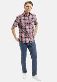 Czerwona i niebieska koszula w kratę z krótkim rękawem, z dwiema kieszeniami na piersi, w połączeniu z niebieskimi dżinsami i białymi trampkami. Model stoi z rękami na biodrach.