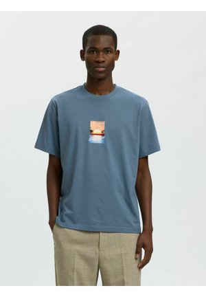 Mann trägt blaues T-Shirt mit Sonnenuntergangs- und Ozeangrafik, kombiniert mit beigen Hosen, steht vor schlichtem grauem Hintergrund.