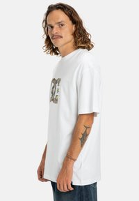 Balta medvilnė t-shirt su trumpomis rankovėmis ir apvaliu iškirpimu. Priekyje yra spalvingas grafiškas logotipas ir laisvas siluetas.