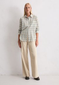 Kobieta stojąca przy białej ścianie, ubrana w beżową koszulę w kratkę zapinaną na guziki, kremowe szerokie spodnie z korduroyu oraz ciemnobrązowe buty.