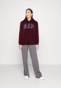 Bordo dukserica s "GAP" logom, kengurska džep, i kapuljača s vezicom; u paru s svijetlosivim rebrastim hlačama širokih nogavica i bijelim cipelama.