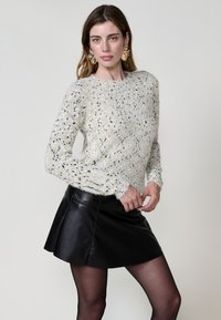 Hellfarbener, strukturierter Pullover mit schwarzen und gelben Sprenkeln, kombiniert mit einem schwarzen Leder-Minirock. Hat lange, leicht ausgestellte Ärmel.