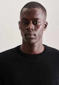 Maglione a coste nere con scollatura tonda. Tessuto liscio, forma aderente e dettagli sottili attorno all'area del collo. Il modello ha i capelli corti.
