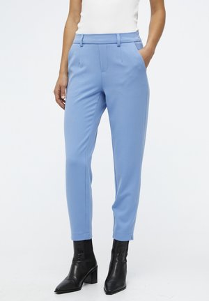 Femme portant un pantalon slim bleu clair avec poches avant et des bottines à talons noires, debout devant un fond blanc.