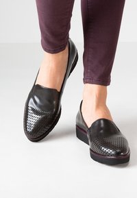 Scarpe nere slip-on con parte anteriore perforata, parte superiore liscia e tacco a trapezio basso, caratterizzate da un accento bordeaux lungo la suola.