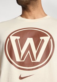 Krémové bavlnené tričko s veľkým hnedým kruhovým logom so výrazným písmenom 'W' uprostred a malým logom Nike dole.