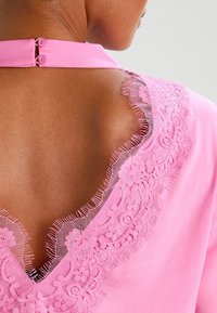 Blusa rosa con scollo a V sul retro rifinito in pizzo, dotata di due bottoni di chiusura al colletto. Texture morbida e dettagli in pizzo delicati.