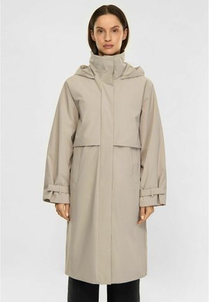 Woman wearing a long beige raincoat with a high collar, hood, and belted cuffs, standing against a plain white background.