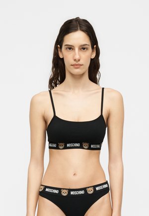 Nő fekete Moschino bralette-et és hozzáillő bugyit visel, amelyeken márkázott plüssmackó logó található az elasztikus pántokon, egyszínű háttér előtt áll.