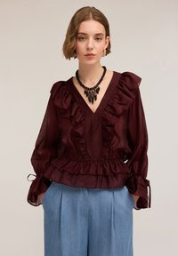 Blusa ruggine con volant e scollatura a V profonda, tessuto trasparente, vita elasticizzata e maniche legate, abbinata a pantaloni azzurri a vita alta.