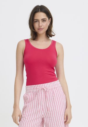 Kobieta o średniej długości brązowych włosach, ubrana w bezrękawnik w czerwonym kolorze i białe spodnie z czerwonymi pionowymi paskami.