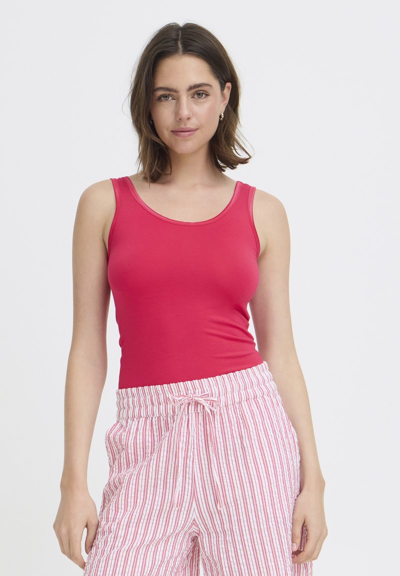 Femme aux cheveux bruns mi-longs portant un haut rouge sans manches et un pantalon blanc avec des rayures verticales rouges.