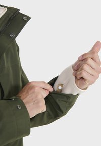 Grüne wasserdichte Jacke mit einem Fleece-Innenfutter. Verfügt über einen Reißverschluss und Druckknopfverschlüsse, mit einem strukturierten Handgelenkbund und strapazierfähigem Stoff.