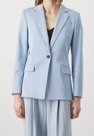 Kvinde iført en lyseblå skræddersyet blazer med en enkelt knap og matchende lyseblå, brede bukser med striber.