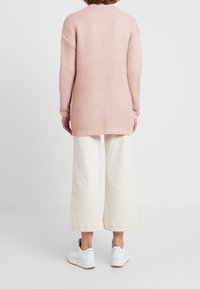 Helle rosa gestrickte Pullover mit strukturierter Oberfläche, langen Ärmeln und lässiger Passform, kombiniert mit cremefarbenen weit geschnittenen Hosen und weißen Sneakers.