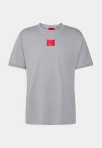 Camiseta de algodón gris con escote redondo, mangas cortas y un parche cuadrado rojo con la inscripción "HUGO" en la parte izquierda del pecho.