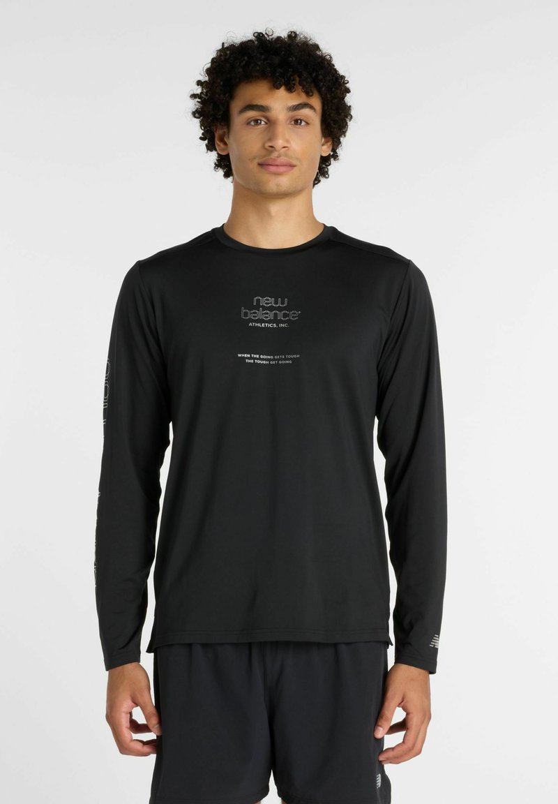 Jeune homme aux cheveux bouclés portant un t-shirt de sport à manches longues noir New Balance et un short noir assorti sur un fond blanc.