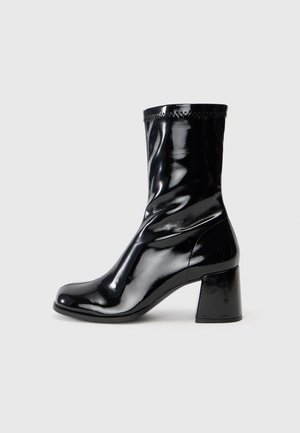 Botte mi-mollet noire en cuir verni brillant avec un bout arrondi et un talon bloc, présentée sur un fond gris clair uni.