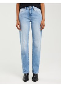 Inimene, kes kannab helesiniseid sirge säärega pükse, musti saapaid ja musta särkiga, käed vabalt külgedel, valgel taustal.