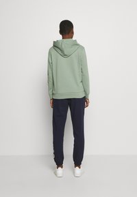 Sudadera con capucha de color verde claro con cordón en la capucha, combinada con pantalones jogger azul marino que presentan un diseño vertical colorido en la pierna izquierda, zapatillas deportivas blancas.