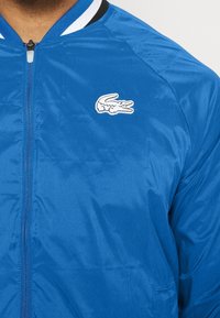 Blå bomberjacka i glänsande tyg, med en svart och vit randig krage och en vit krokodil-logotyp på bröstet. Dragkedja fram.