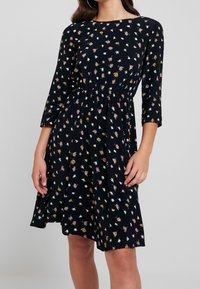 Schwarzes Kleid mit drei Viertelärmeln, das ein florales Muster in Pink, Gelb und Weiß aufweist. Elastischer Taillenbund, knielang, lockere Passform.