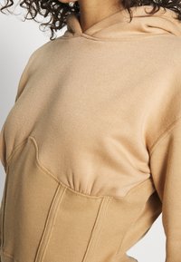 Sudadera beige con capucha, con ribetes en el dobladillo y detalles en el cuerpo. Presenta una textura suave y pliegues en la parte delantera para un corte más ajustado.